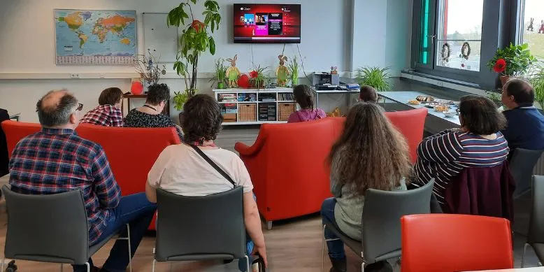 Mehrere Menschen sitzen im Clubhaus vorm Fernseher