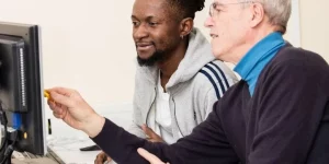 Zwei Männder sitzen am PC, der Ältere erklärt dem jüngeren etwas. Der jüngere Mann hat einen Mgrationshintergrund.