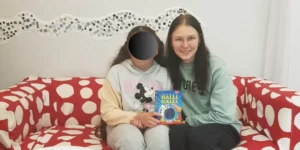 Lernpatin sitzt mit ihrem Mentee auf einen roten Sofa mit weißen Punkten. Sie halten ein Spiel in der Hand.